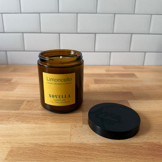 An amber glass candle jar with a yellow label reading 'Limoncello' and 'NOVELLA' sits on a wooden countertop with its black lid placed beside it. A white subway tile backsplash is visible in the background.